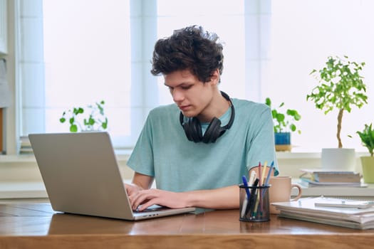 Young male college student sitting at desk at home using laptop. Guy 19-20 years old studying at home, using a computer for education leisure communication