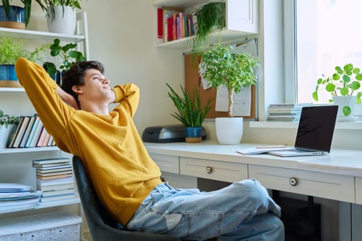 Young male calm college student relaxing on chair at home, smiling relaxed guy taking break between online classes, laptop notebook on desk. E-learning, education, youth concept