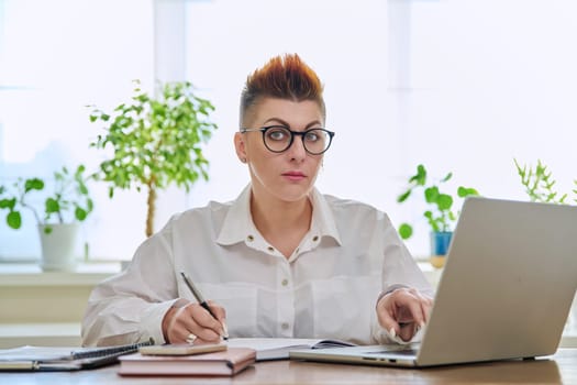 Portrait of mature businesswoman working at home on computer laptop. Smiling middle-aged female looking at camera sitting at desk in home office. Remote business work, career, management, marketing