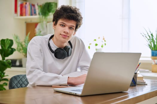 Young college student guy sitting at desk with laptop computer looking at camera. Handsome curly young male wearing headphones in home interior. Education, lifestyle, youth 19,20 years old