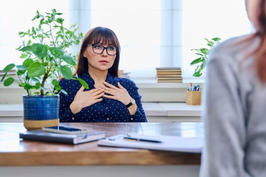 Mature sad woman in therapy session with mental professional psychologist, sitting at table in office of therapist counselor social worker. Psychology psychotherapy mental assistance treatment support