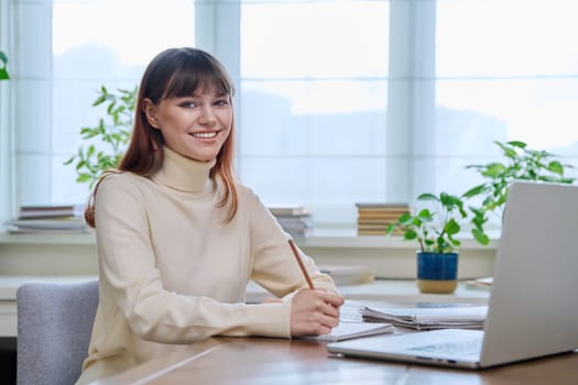 Young female college student studying at home at desk using computer laptop, writing in notebook, smiling looking at camera. E-learning, education, technology, knowledge, youth concept