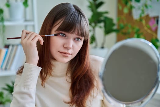 Young woman with cosmetic products on table, doing makeup looking in mirror at home. Cosmetics, fashion, youth, lifestyle, beauty concept