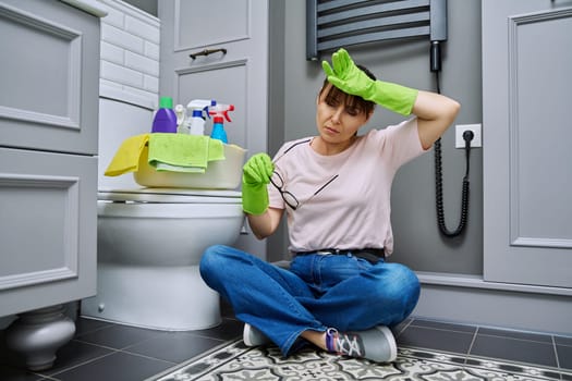 Tired woman sitting on the floor after cleaning bathroom, toilet. Routine house cleaning, home hygiene, housecleaning service, housekeeping, housework