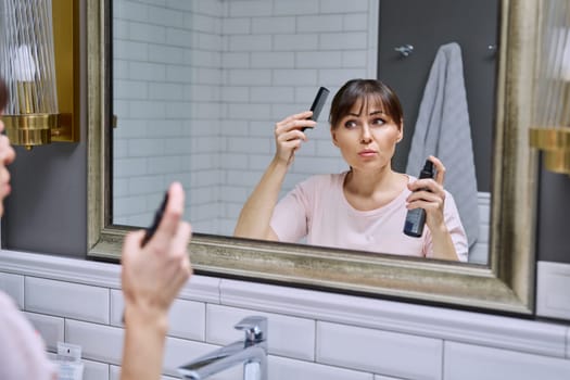 Middle-aged woman with setting spray for styling hair fixation comb looking in mirror, doing hair styling, in bathroom. Beauty, age, cosmetics, hair accessories