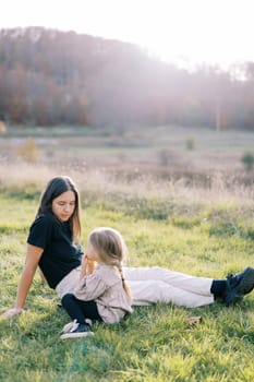 Mom sits on the green lawn and looks at her little daughter sitting next to her. High quality photo