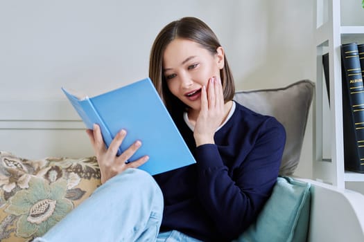 Young woman reading paper book, relaxed sitting on sofa at home. Fiction, hobby, leisure, youth concept