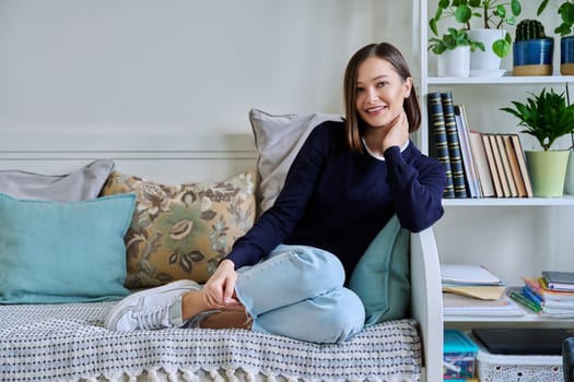 Portrait of young attractive smiling woman in home interior. Happy cheerful relaxed 20s female looking at camera, sitting on couch. Beauty, youth, happiness, health, lifestyle concept