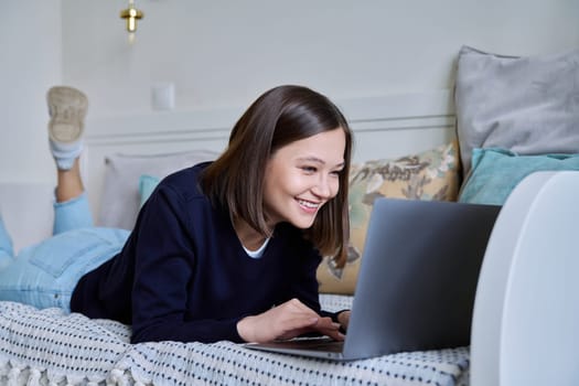 Young woman using laptop computer, typing on keyboard, lying on sofa at home. Internet online technologies for work communication study leisure