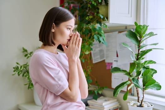 Profile view close-up of young woman's face with hands folded in prayer, dreams, memories. Lifestyle, feelings, mental physical health, loneliness, stress, depression, difficulties, youth concept