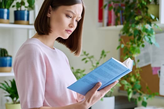 Serious concentrated young woman reading book at home near window. Important study literature guide, interesting fiction, hobby leisure reading