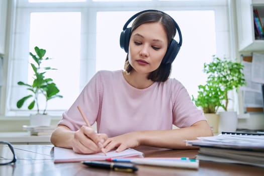 Webcam portrait of young 20s female student wearing headphones, sitting at home at desk, listening writing. Internet online webinar, video chat call conference, technology education training