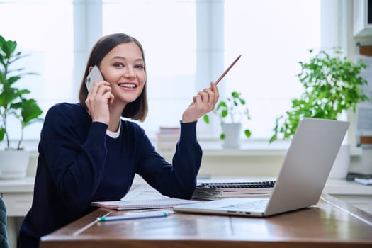 Happy smiling young woman sitting at desk at home office with computer laptop and talking on smartphone. Female working, studying remotely, freelancer, teacher, student