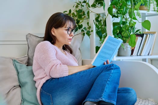 Mature woman reading paper book sitting on couch at home. Relaxation, literature hobby, fiction, lifestyle, people concept