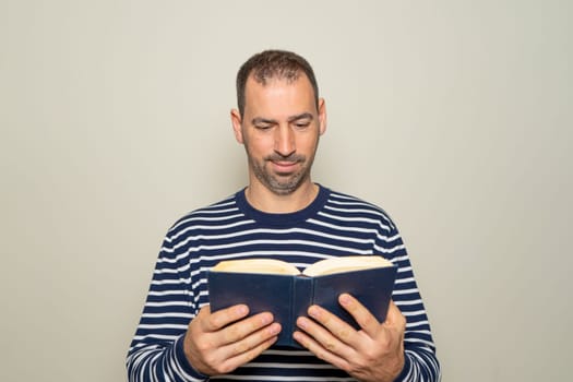 Hispanic man with a beard in his 40s wearing a striped sweater reading a paper book, he is focused and excited about the magnificent content of the story. Isolated on beige background