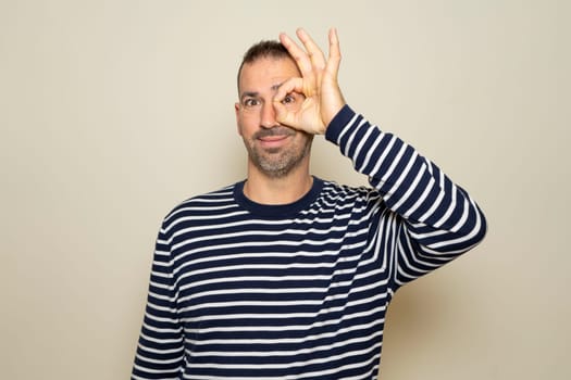 Bearded Hispanic man in his 40s wearing a striped sweater smiling while making an ok gesture over his eye in approval, isolated on beige studio background