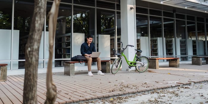 Young businessman working online with laptop while sitting on bench outside of office building.