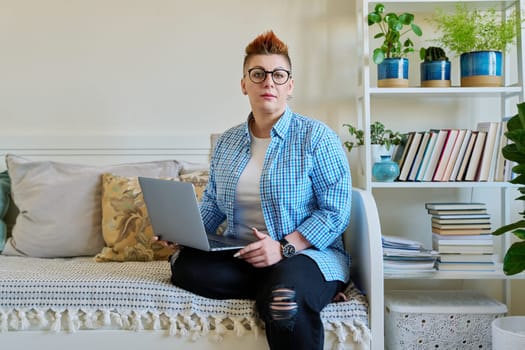 Middle aged woman sitting on couch at home using laptop, looking at camera. Serious female with red haircut in casual clothes working on computer, relaxing, communicating