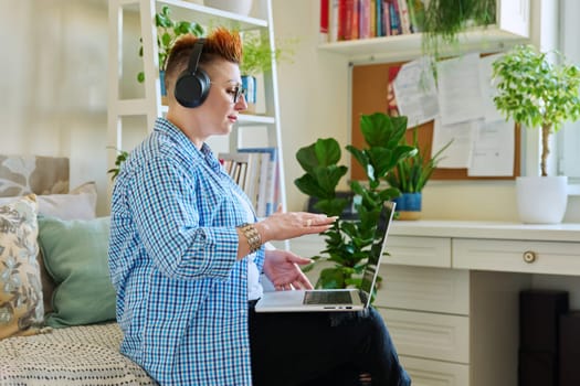 Middle aged woman freelancer in headphones using laptop computer sitting on couch at home. 40s female looking at screen video chat conference call online meeting training mentoring technology for work