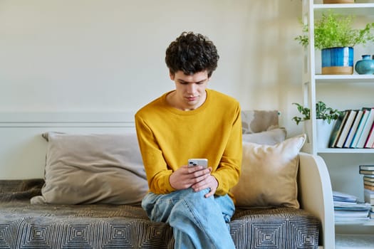 Young guy holding smartphone in hands sitting on couch at home. Handsome university college student 19-20 years old in yellow with curly hair. Technologies, mobile apps, study communication leisure