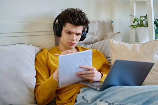 Young guy college student in headphones using laptop making notes, sitting on couch at home. Webinar listening audio online conference chat lesson e-learning education remote classes internet meetings