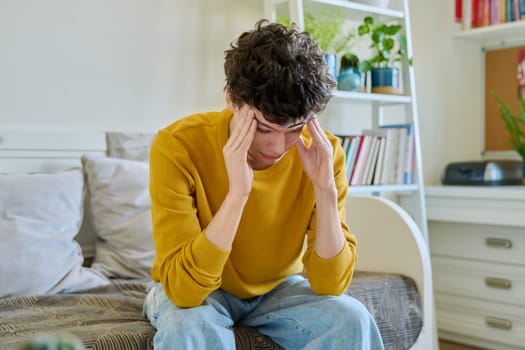 Sad upset tired young man sitting on couch at home, touching his head with hands. Health problems, headaches, troubles, difficulties in study, family relationships, mental health, stress, depression