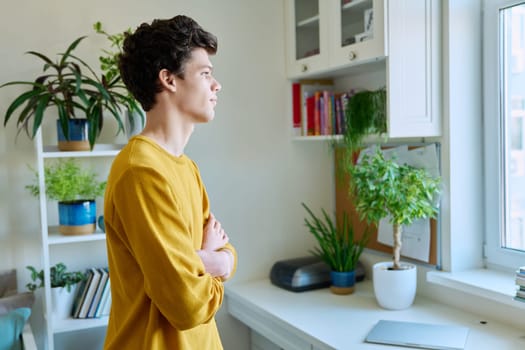 Profile portrait of young handsome guy with crossed arms, in home interior. Confident serious male 19-20 years old in casual yellow with curly hairstyle looking at window. Lifestyle, youth concept