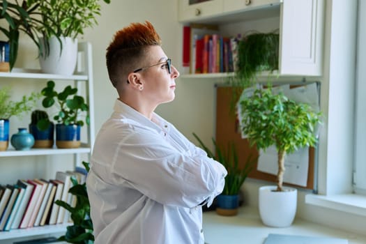 Profile portrait of serious confident middle-aged woman in glasses with red haircut. Female with crossed arms looking at window in home interior. Mature people, lifestyle, health, life concept