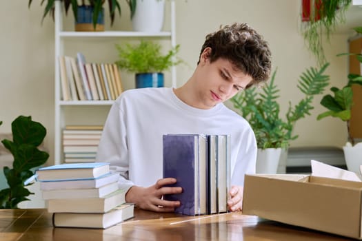 Satisfied young male customer buyer sitting at home unpacking cardboard box parcel with new books, online purchases. Delivery by mail, internet store bookstore