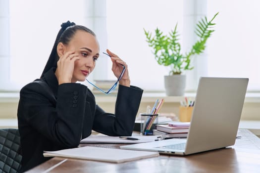 Tired sad business woman at the workplace. Upset female sitting at desk with laptop computer, experiencing stress pain headache. Business, remote work, health problems, freelance concept