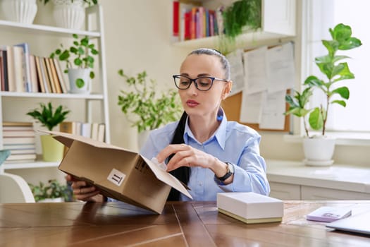 Satisfied female customer buyer sitting at home unpacking cardboard box with online purchases. Woman unpacking boxes with cosmetics care products vitamins perfumes. Delivery by mail, online store