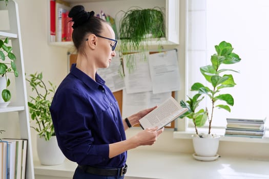 Middle-aged woman reading a fiction book at home, at home near the window, winter season