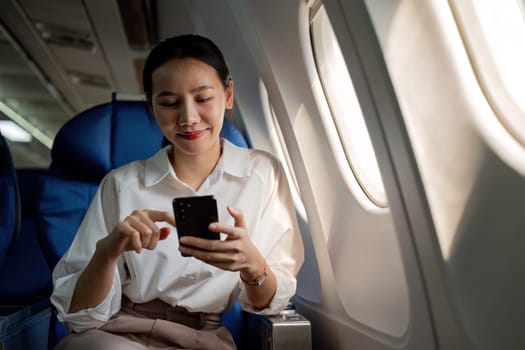 Young Asian woman uses mobile phone to check news information Sitting near the window in business class, airplane class during flight, travel and business concept.