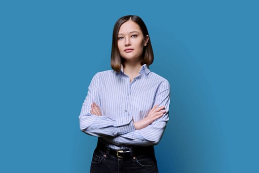 Young confident serious woman with crossed arms on white blue background. Business female in shirt, student worker owner entrepreneur looking at camera. Business, work, education, people concept