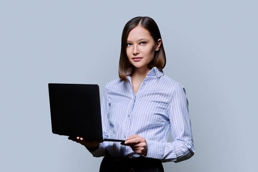 Serious young business woman with laptop on grey background. Confident successful female holding computer in hands looking at camera. Business, work, training, education, modern digital technologies