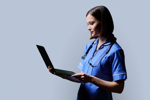 Serious young female nurse using laptop computer, profile view on gray studio background. Mobile apps applications technologies in medical services health professional assistance medical care concept