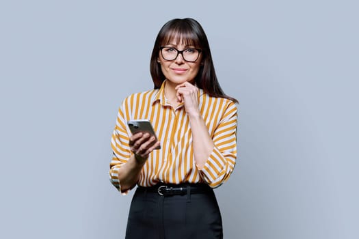 Middle aged smiling woman holding smartphone on grey background. Mature female looking at camera with phone in hands. Technologies mobile applications internet work business leisure communication