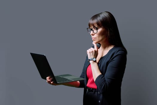 Serious middle aged woman holding laptop on gray studio background. Confident mature female working using computer. Internet online technology, business, work, service, lifestyle