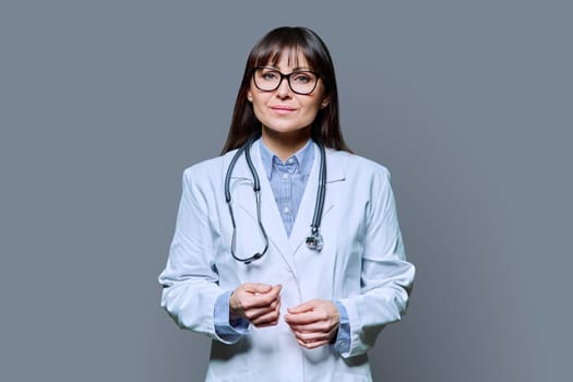 Confident friendly smiling middle-aged female doctor in white lab coat with stethoscope, looking at camera on gray studio background. Healthcare, medicine, staff, treatment, medical services concept