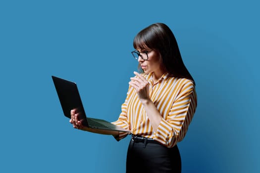 Serious middle aged woman holding laptop on blue studio background. Confident mature female working using computer. Internet online technology, business, work, service, lifestyle