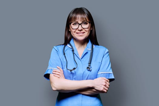 Portrait of smiling middle-aged woman nurse in blue with crossed arms with stethoscope on grey studio background. Medical services, nhs, health, professional assistance, medical care concept