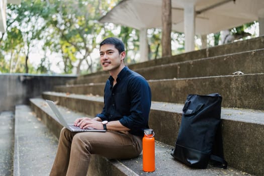 Young businessman working online with laptop while sitting on bench outside of office building.