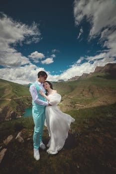 Young newlyweds, bride and groom hugging with a beautiful view of the mountains, blue sky with white clouds and green meadows in the background.