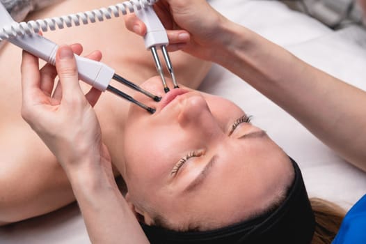 Close-up of a professional cosmetologist's hands touching a woman's cheek with equipment. Young woman lying and relaxing during massage.