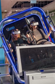 Moscow, Russia, March 14, 2024. Two woman is sitting in an electric blue virtual reality chair, wearing a headset. The machine combines engineering and fun in a display device