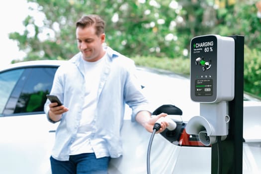 Modern eco-friendly man recharging electric vehicle from EV charging station, using Innovative EV technology utilization for tracking energy usage to optimize battery charging on smartphone.Synchronos