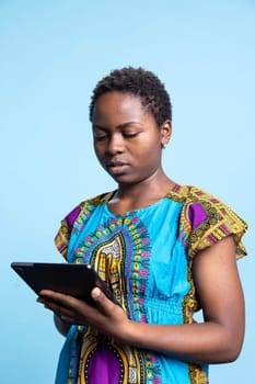 African american woman is using a modern tablet in studio, scrolling through online webpages over blue background. Girl holding device to stay connected with people on social media platforms.