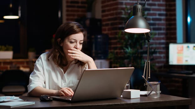 Weary accountant struggling to remain awake at workspace desk while inputting data on laptop, panning shot. Close up of exhausted businesswoman yawning in office while working overnight, camera B