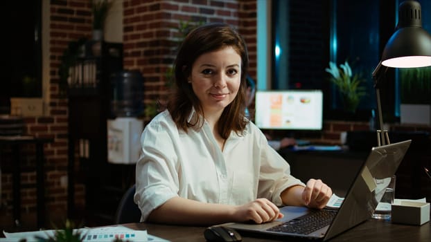 Portrait of smiling accountant researching key data for company project, doing tasks in office overnight. Happy employee sitting at desk in front of laptop, looking over accounting figures, camera A