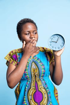 Surprised woman looking at blue round clock to check the time, trying to meet deadline using timepiece object over blue background. African american person holding watch decoration.